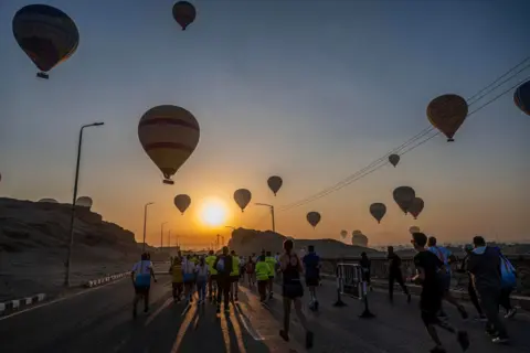 Khaled DESOUKI / AFP The sun rises in the sky filled with hot air balloons as people run along a road as part of the Egyptian Marathon.