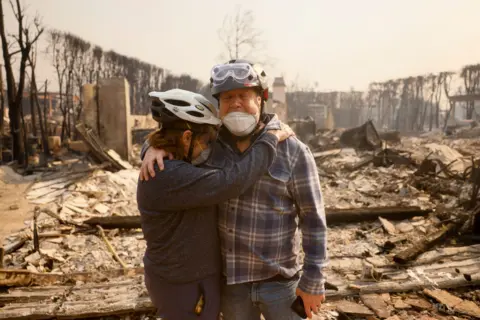 Etienne Laurent/AP A husband and wife embrace in their fire-ravaged neighborhood, in front of burnt rubble, both wearing helmets and protective face masks, after the Palisades Fire swept through in the Pacific Palisades neighborhood of Los Angeles on Wednesday.