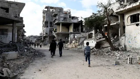 Reuters People, including a child on a crutch, missing the lower half of his left leg, walk past the grey, dusty rubble of houses and buildings destroyed during the war in Al-Bureij in central Gaza
