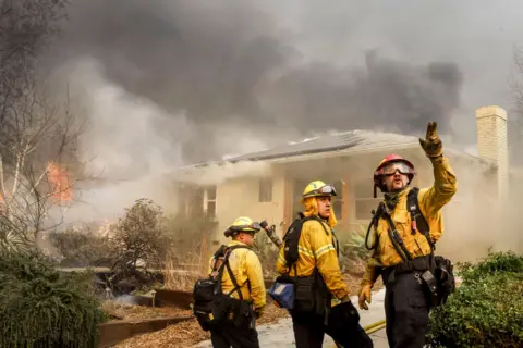 Caroline Brehman/EPA Three Los Angeles County firefighters look away from camera and gesture next to a burning home while trying to protect homes from the Eaton wildfire in Altadena on Wednesday