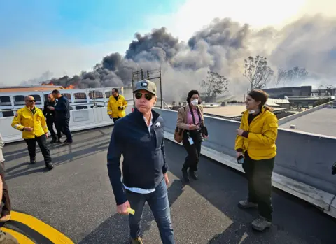 Jeff Gritchen/MediaNews Group/Orange County Register California Governor Gavin Newsom looks away from camera as he surveys damage during the Palisades Fire, surrounded by personnel and with smoke rising in the background.