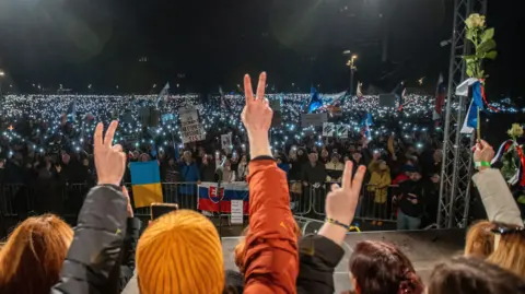 EPA An evening protest in Bratislava, where several demonstrators with their backs to the camera give victory salutes and one holds up a rose. They face a much larger group of protesters behind barriers holding banners and Slovakian and Ukrainian flags