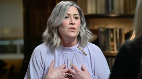 Jeff Overs/BBC Caroline Darian gesticulates in a picture taken during her interview, bringing her hands up to her chest. She is a woman in her 40s with her hair in a bob, wearing gold jewellery.