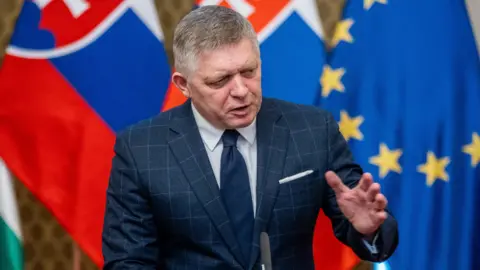 EPA Robert Fico, wearing a blue checked suit and blue tie, gestures with his left hand as he holds a press conference on 21 January. Slovak and EU flags stand in the background