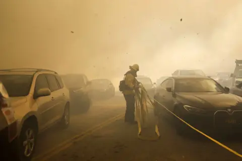 Caroline Brehman/EPA A Los Angeles firefighter drags a hose past cars trying in a smoky street in Pacific Palisades.