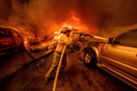 Ethan Swope/AP Flames can be seen in the background as a firefighter lifts a water hose to spray a burning vehicle