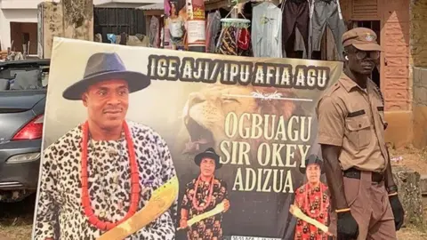 Adaobi Tricia Nwaubani A uniformed security officer stands to the right of a poster advertising a ceremony for a man called Sir Okey Adizua to become a 'Leopard Slayer'. There are three images of a man holding a gold sword, wearing a large red bead necklace and a wide-brimmed black hat