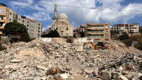 Scarlett Barter / BBC Rubble of the apartment block in the foreground, and in the background a few apartment blocks of various styles, flanking a mosque. A yellow digger picks through the detritus.